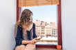 © Kristina Blokhin - Young woman looking at phone sitting on window sill of apartment with view of old market square in town of Warsaw, Poland