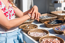 Young Woman Shopping For Colorful Stone Beach Bead Bracelets Trying On Jewelry In Outdoor Market Shop Store In European, Greece, Italy Or Mediterranean Town Village In Summer