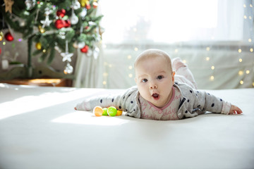 Wall Mural - Surprised baby girl lying down on bed at home. Christmas tree in background.