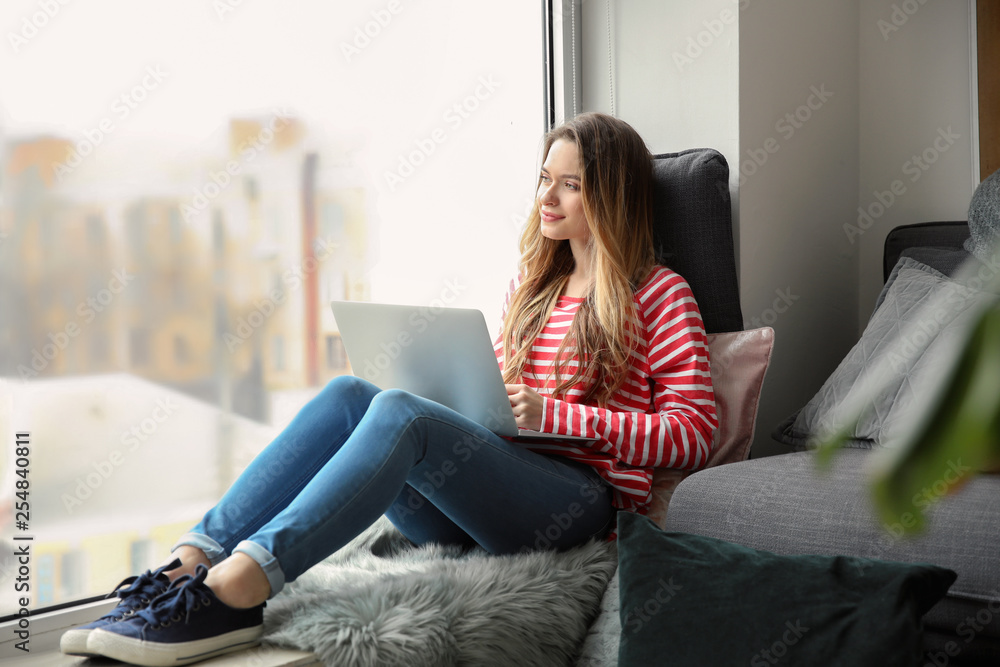 Beautiful young woman with laptop sitting on window sill