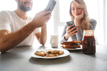 Wall Mural - Loving couple sitting at the kitchen have a breakfast using mobile phones.