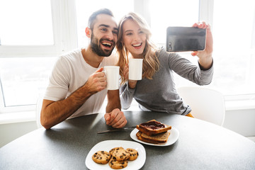 Wall Mural - Young loving couple sitting at the kitchen have a breakfast take a selfie by mobile phone.