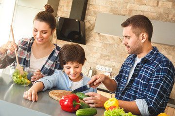 Wall Mural - Family at home standing in kitchen together father showing son cutting vegetables while mother mixing salad happy