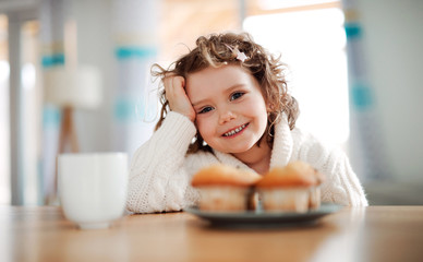 Wall Mural - A portrait of small girl sitting at the table at home, eating muffins.