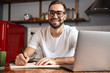 © Drobot Dean - Photo of smiling man writing down notes while using silver laptop on kitchen table