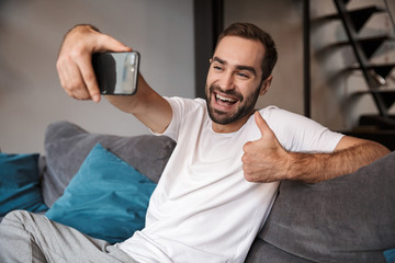 Sticker - Photo of joyous bachelor holding and taking selfie on cell phone while sitting on couch in living room