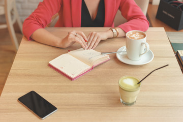 A woman is sitting at a table in a cafe with a spare book. On the table stands a mug of cappuccino, a telephone and a matcha tea. Negotiation concept in co working place. Living coral concept