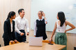 A diverse team stand around a laptop for a discussion in a meeting room in the office during the day. They are ethnically diverse and includes a Caucasian, Indian and Eurasian woman and a Chinese man.