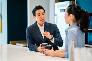 An Asian Chinese man in a suit having a discussion with his Asian woman colleague in an office during the day. They are both holding mugs of a hot beverage (coffee or tea) and talking.