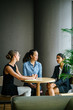 © Danon - A diverse group of three young women sit around a table and smile while having a casual discussion. They're dressed professionally and are in an office meeting room.