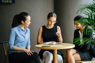 A young Indian Asian woman is having a business meeting with her team in a conference room. Her colleagues are diverse composed of a Caucasian brunette woman and a Chinese Asian woman