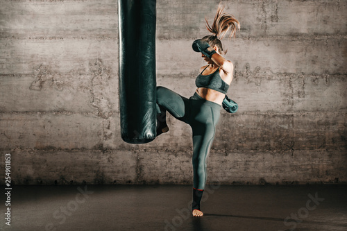 Dedicated strong brunette with ponytail, in sportswear, bare foot and with boxing gloves kicking sack in gym Фотошпалери