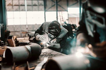 Iron worker in protective suit, mask and gloves welding pipe. Workshop interior.