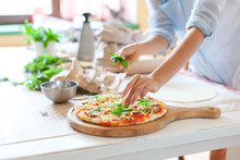 Woman Is Cooking Pizza In Cozy Home Kitchen. Female Hands Are Decorating Italian Dinner With Greens, Fresh Basil, Arugula. Homemade Pizza Is Served On Wooden Board On Table. Lifestyle Moment. Close Up