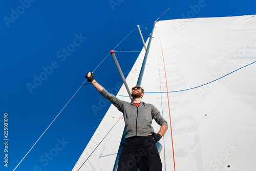 Fotografía Yacht captain with a beard stands on sail boom on a sailing yacht, holding the rope in his hand and smiling, feeling happy