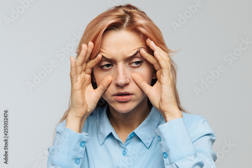 Very tired young woman in blue shirt wants to sleep at work or university, opening her eyes with the fingers, squinting so as not to fall asleep. Lack of sleep, overwork, boredom, wide open eyes