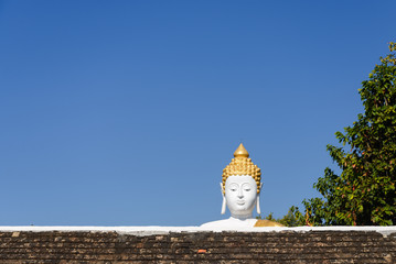 Outdoor sunny view of Buddha statue's head stand out over old rough clay roof of temple in Chiang Mai, Thailand. Traditional asian big buddha statue taller than temple.