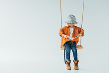Cute Kid In Jeans And Orange Shirt Sitting On Swing And Reading Book On Grey Background