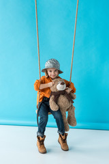 Wall Mural - cute kid sitting on swing and holding teddy bear on blue background