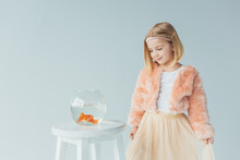Adorable Kid In Faux Fur Coat And Skirt Looking At Fishbowl On Stool Isolated On Grey