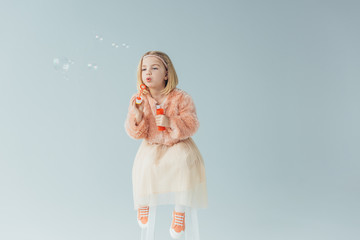 adorable kid in faux fur coat and skirt sitting on highchair and blowing soap bubbles isolated on gr