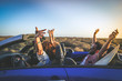 Side view group three euphoric friends in convertible car twisting and waving Two curly girls and charming bearded man on vacation having fun in topless auto Arms up palms open driving in the sunlight