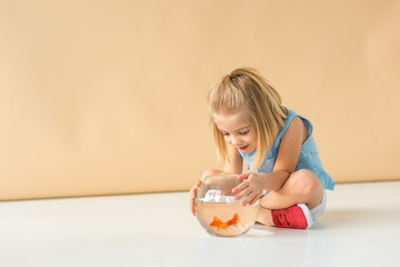 Wall Mural - adorable kid sitting with crossed legs and looking at fishbowl on beige background