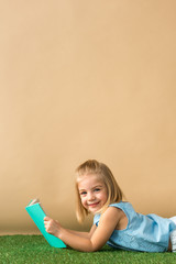 Wall Mural - smiling and cute kid lying on grass rug and holding book on beige background