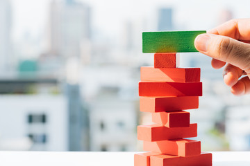 Businessman holding green block on red tower stack from wooden blocks toy with city and sky backgrounds. Business strategy, learning and development concepts.