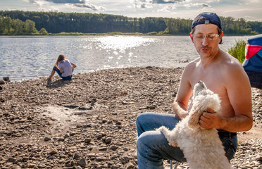 summer  landscape  white man in jeans and cap sunbathing on the shore of the grill. next to the white dog. a teenage girl in a white t-shirt sitting on the river Bank.