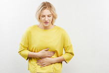 Woman With Suffer Expression On Face Holding Stomach And Squirming With Pain. Indigestion, Intestinal Problems, Gas And Bloating On A White Background