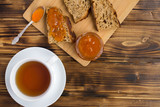 Fototapeta Kuchnia - Sliced bread with apricot jam on the cutting board and tea in the white cup on the brown wooden background.Top view.Copy space.