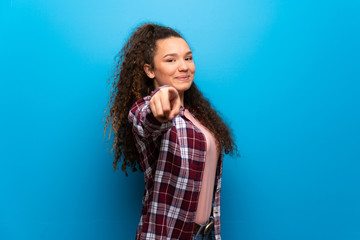 Teenager girl over blue wall points finger at you with a confident expression