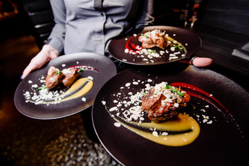 The girl waiter carries three plates of food. Meat steak garnished with two sauces on a dark plate. Close-up. Wide angle photography. 