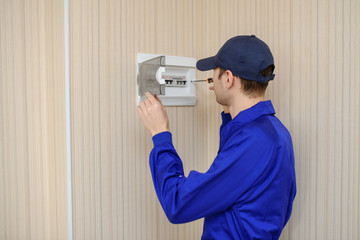 Wall Mural - lateral view of a young eletrician in blue overall disassembling a electrical panel with fuses in a house.