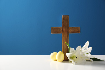 Wooden cross, Easter eggs and blossom lilies on table against color background, space for text