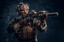 Elite Unit, Special Forces Soldier In Camouflage Uniform Holding An Assault Rifle With A Laser Sight And Aims At The Target. Studio Photo Against A Dark Textured Wall