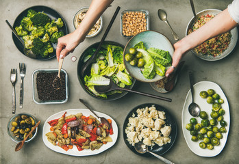 Vegan dinner table setting. Healthy dishes in plates on table. Flat-lay of vegetable salads, legumes, beans, olives, sprouts, hummus and woman hands mixing ingredients on plate, top view