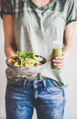 Wall Mural - Healthy dinner or lunch. Woman in t-shirt and jeans standing and holding vegan superbowl or Buddha bowl with hummus, vegetable, fresh salad, beans, couscous and avocado and smoothie in hands