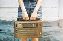 Hipster Girl In Denim Jeans Shorts Is Holding Retro Radio In Hands. Style Concept. Vintage Music On Wooden Background. Modern Technologies Outdoors.