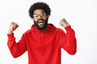 © Cookie Studio - Guy feeling awesome, winning, celebrating victory raising fists in triumph and success smiling broadly as in good mood, standing upbeat in red hoodie over gray background, achieving great result