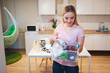© estradaanton - Recycling. Young smiling woman holding electronic waste in the container with green recycle icon on kitchen background