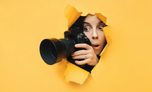 A Paparazzi Funny Girl With A Camera Looks Out From Cover And Looks At What Is Happening With Her Mouth Open. Yellow Paper, Torn Hole. Tabloid Press. In Search Of The Plot For Photo Stocks.