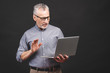 © denis_vermenko - Using skype. Senior aged bearded old man in eyeglasses holding laptop computer isolated against black background.