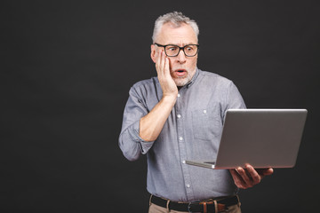 Wall Mural - OMG! I'm shocked! Scared senior aged bearded old man in eyeglasses holding laptop computer isolated against black background.