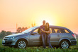 © bilanol - Young couple slim attractive long-haired woman in jeans and t-shirt and handsome sportive man stand embraced leaning on silver car on clear bright sky at sunset or sunrise copy space background.