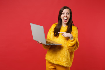 Excited young woman in yellow fur sweater keeping mouth wide open pointing index finger on laptop pc computer isolated on red background. People sincere emotions lifestyle concept. Mock up copy space.