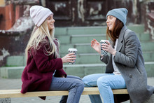 Outdoors Fashion Portrait Of Two Young Beautiful Girls Drinking Coffee. Coffee To Go. Two Friends Relaxing And Drinking Coffee .Coffee Break.Coffee To Go Stylish Hipster Girl Drinking Coffee In Street