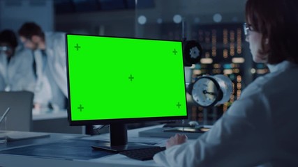 Over the Shoulder Shot: Female IT Scientist Uses Green Mock-up Screen Computer. In the Background Technology Research And Development Laboratory with Scientists, Engineers Working on High Tech Design