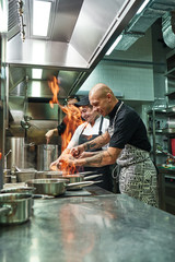 Wall Mural - Very hot Cheerful chef and two his assistants preparing the dish on the stove with an open fire in the kitchen of the restaurant.